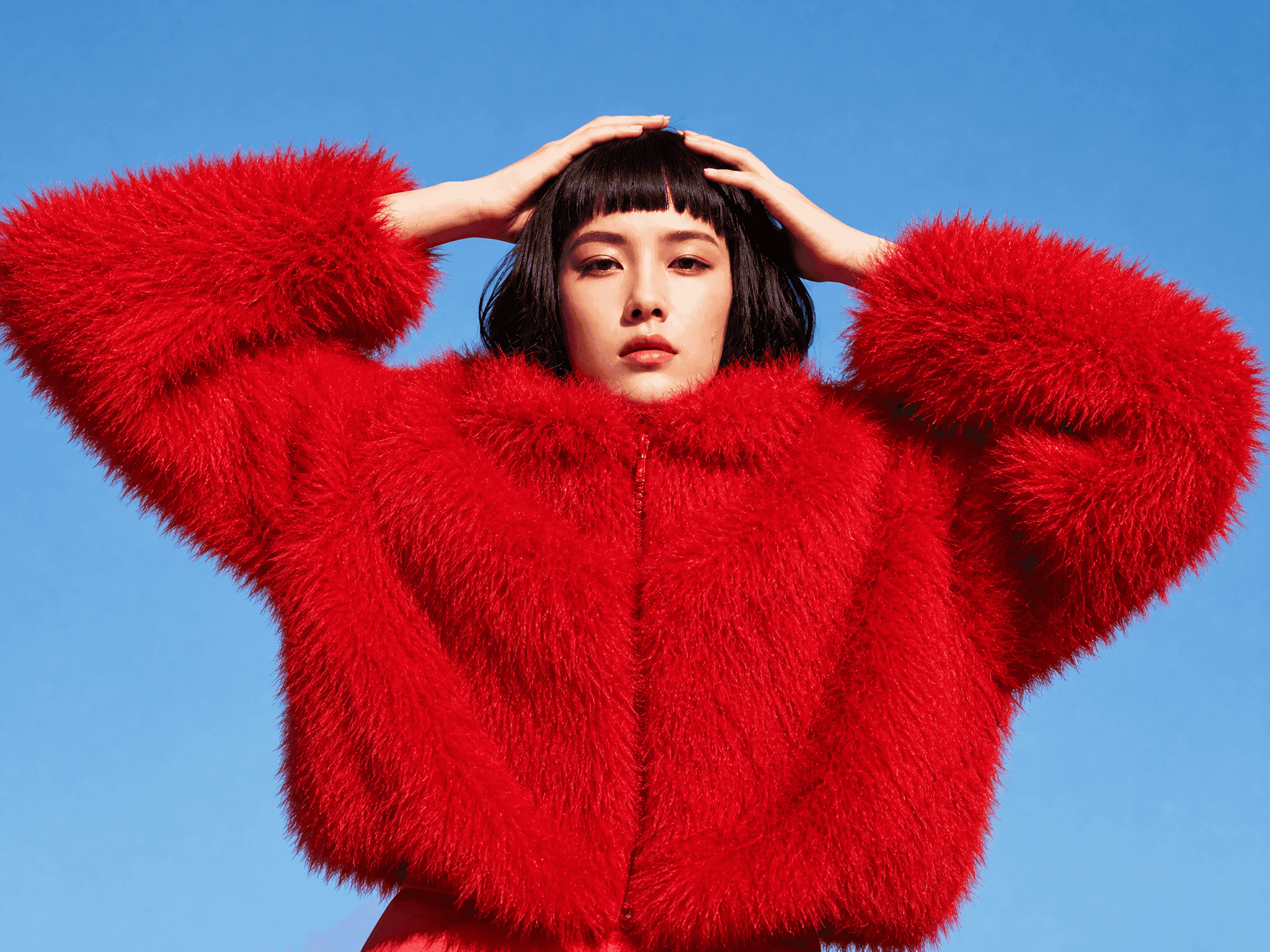 Woman in a red fur jacket with her hands on her head against a blue sky